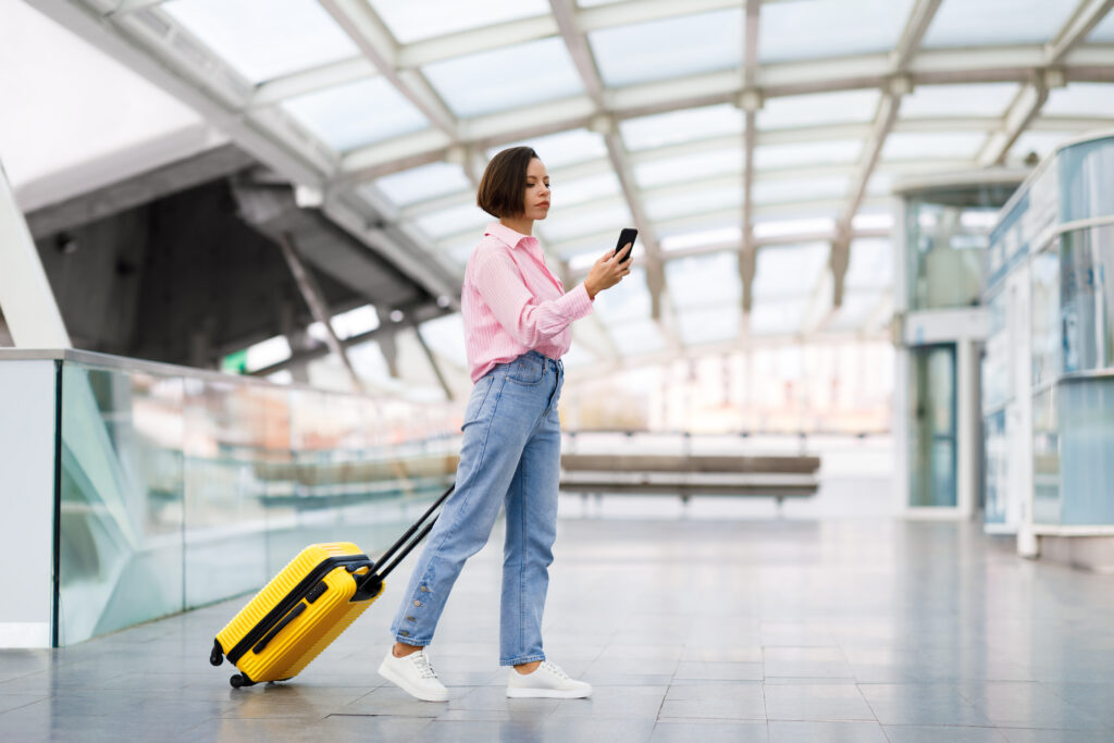 Eine schöne junge Frau mit einem Koffer geht mit ihrem Handy am Flughafen entlang.